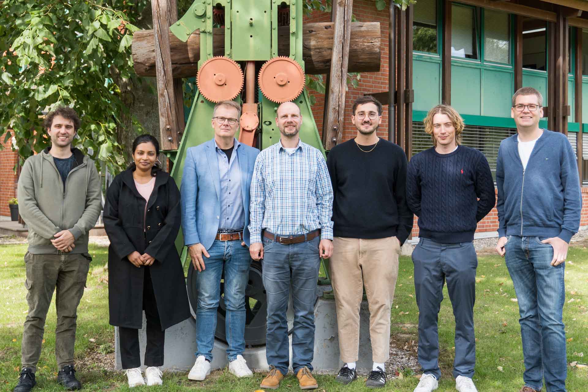Das Team der Machbarkeitsstudie von KI.SH und TimberTec.. Insgesamt 7 Personen sehen in einer Reihe vor einem metallischen Gebilde.